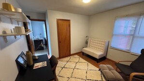 Una sala de estar con un sofá, una silla, una mesa y una alfombra. Se observa una puerta y una ventana con persianas.