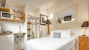Et soveværelse med en seng, et trækommode, et skrivebord med lampe, en mikrobølgeovn og en reol med service.