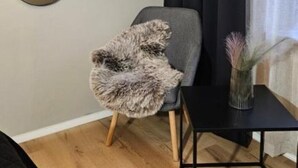 A room with a wooden floor, a grey armchair with a furry throw, a black side table with a vase, and a large mirror on the wall. - Independent apartment with kitchen (Norrbottens län)