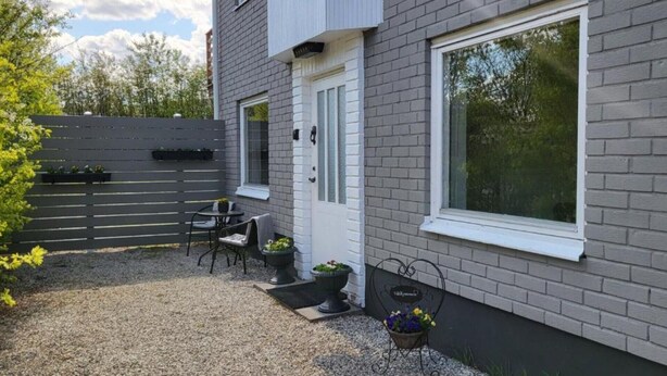 A modern house with a grey brick exterior, a white door, and a large window. There is a patio with a table and chairs, and a small garden area with potted plants. - Independent apartment with kitchen (Norrbottens län)