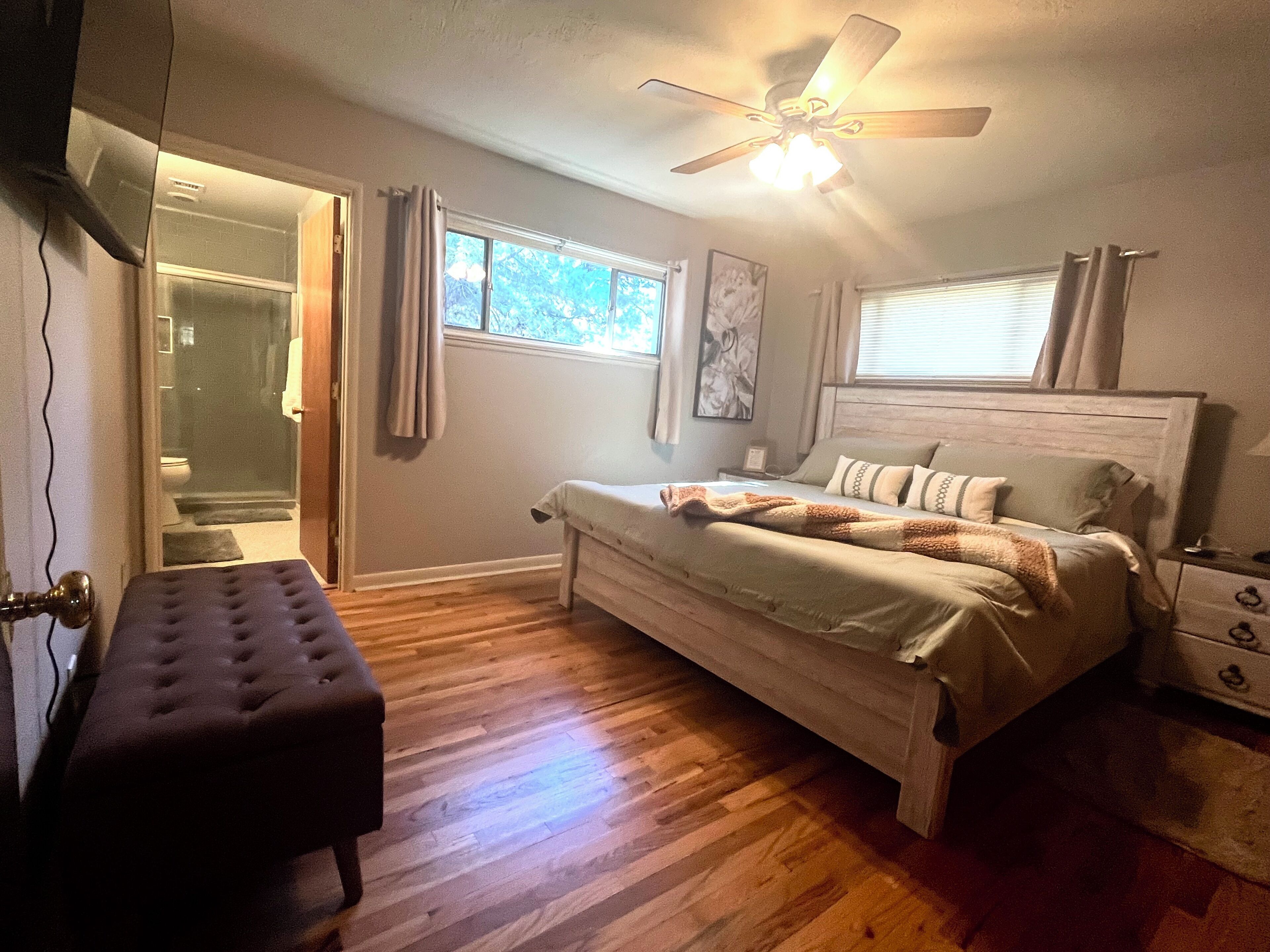 A bedroom with a wooden floor, a bed with a headboard, a bench, a ceiling fan, and a window with curtains.