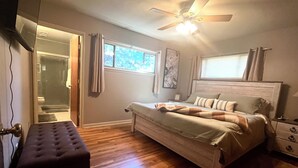 Un dormitorio con suelo de madera, una cama con cabecera, una banca, un ventilador de techo y una ventana con cortinas.