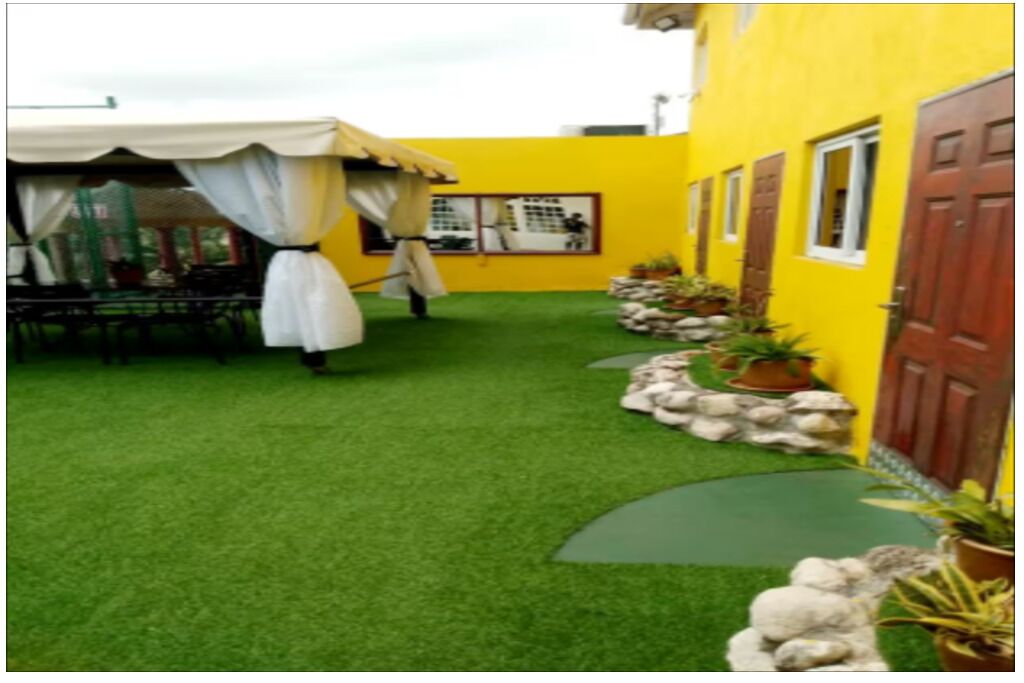A courtyard with artificial grass, a dining area with a table and chairs, and a yellow building with windows and a door.