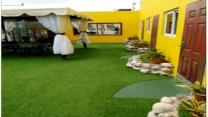 A courtyard with artificial grass, a dining area with a table and chairs, and a yellow building with windows and a door.