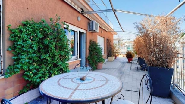 Un patio con una mesa redonda y sillas, rodeado de plantas en macetas y un edificio de fachada roja.