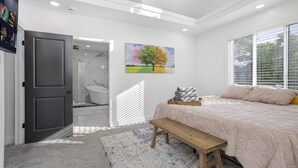 A bedroom with a bed, a bench, a painting, and a bathroom visible through an open door. - Columbus: Victorian Villa (Columbus)