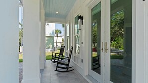 A covered porch with two rocking chairs, a tiled floor, and a glass door leading to a garden. - Lagniappe at Blue Mountain by Avantstay 4BR Coastal Home + Walk to the Beach Community Pool + Gym (Santa Rosa Beach)