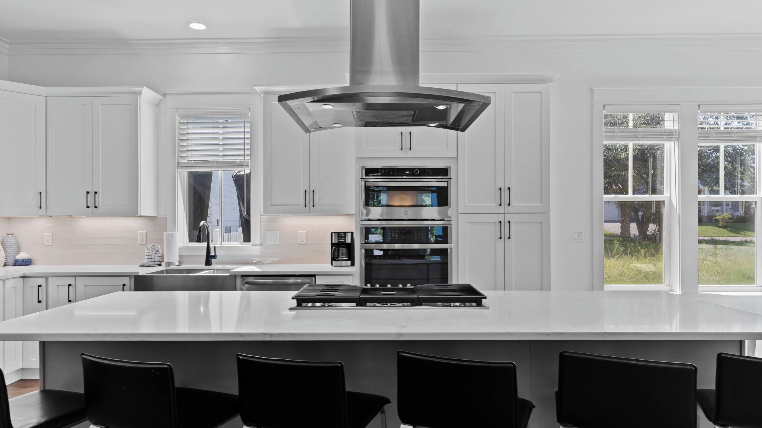 A modern kitchen with white cabinetry, a central island with bar stools, built-in ovens, and a large window with a view of greenery.