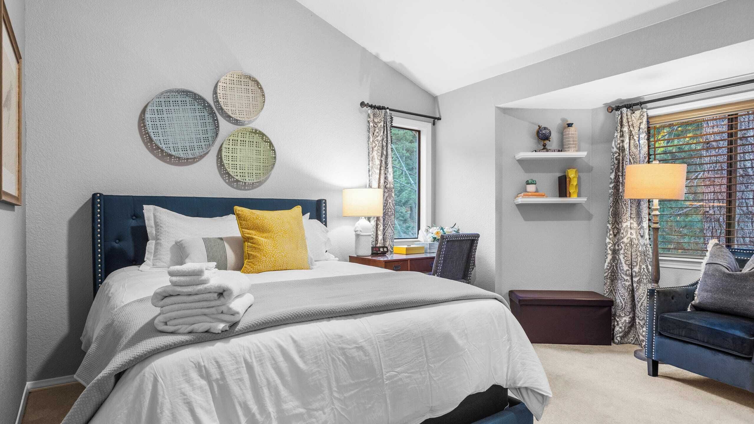 A hotel room with a large bed, a nightstand, a dresser, a window with curtains, and a view of trees.