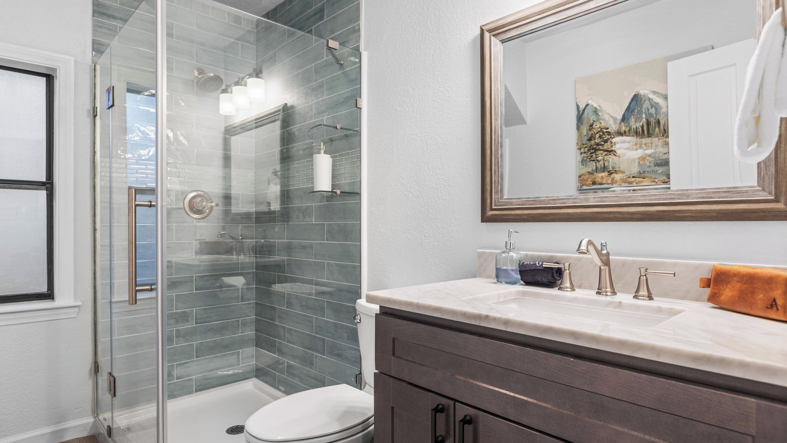 A modern bathroom with a glass shower enclosure, a white toilet, a vanity with a mirror, and a painting on the wall.