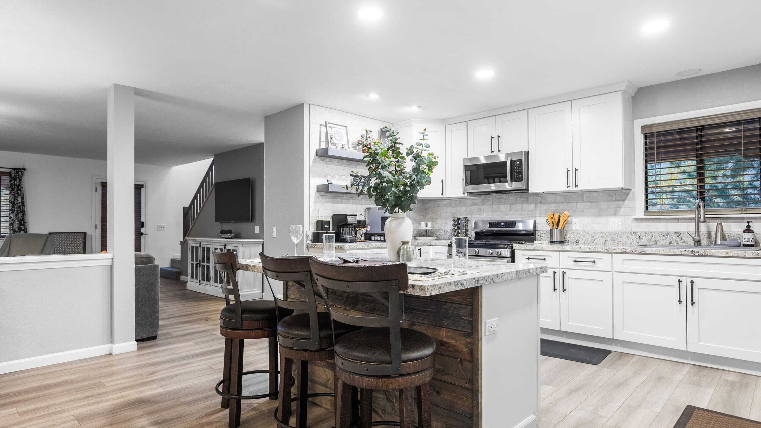 Una cocina moderna con armarios blancos, una isla central con taburetes de bar, electrodomésticos de acero inoxidable y un amplio ventanal con persianas.