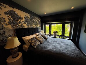 Un dormitorio con una cama grande, una mesita de noche, una lámpara y una ventana con vistas a zonas verdes.