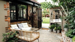 A cozy outdoor seating area with wicker furniture and a brick wall, surrounded by potted plants and a garden. - Balancyoga Guesthouse (West Kingsdown)