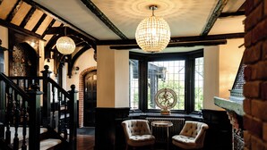 A grand staircase with a chandelier, a fireplace, and two armchairs in a room with exposed brick and wooden beams. - Balancyoga Guesthouse (West Kingsdown)
