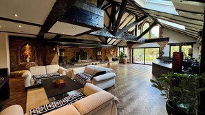 A spacious living area with a high ceiling, exposed beams, a fireplace, and a large window offering a view of the outdoors. - Balancyoga Guesthouse (West Kingsdown)