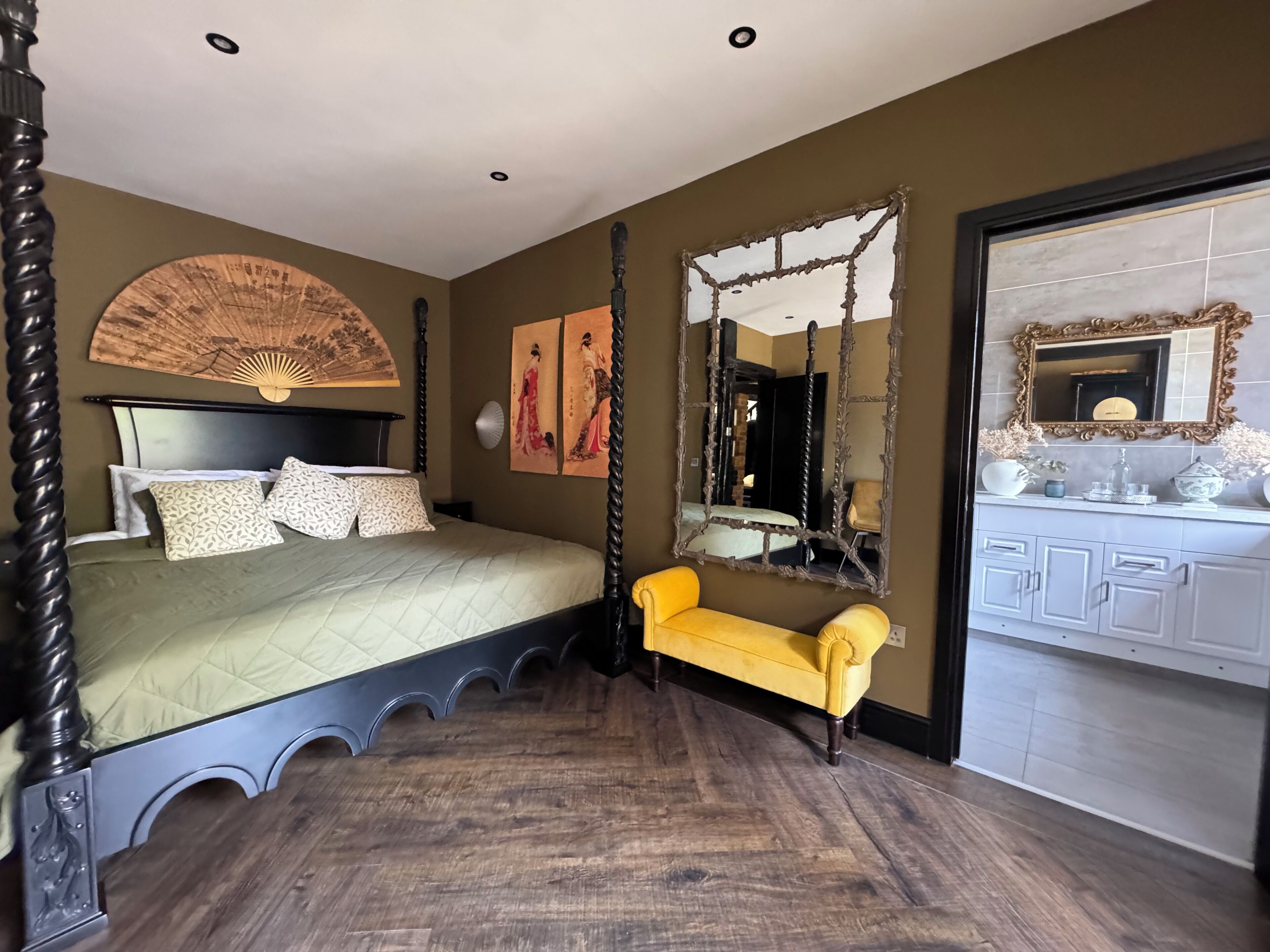 A bedroom with a bed, a yellow chair, a large mirror, and artwork on the walls.