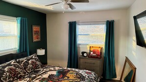 A bedroom with a bed, a ceiling fan, a window with curtains, a bedside table with a lamp, a framed picture, and a suitcase with books. - Charming 1-bedroom cottage in Covington with AC (Covington)