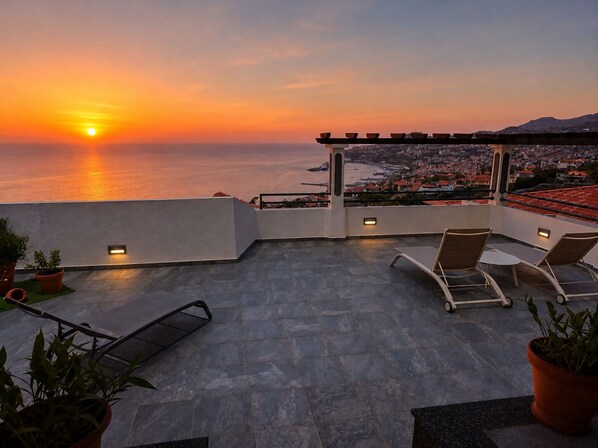 A rooftop terrace with a sunset view, lounge chairs, potted plants, and a pergola.