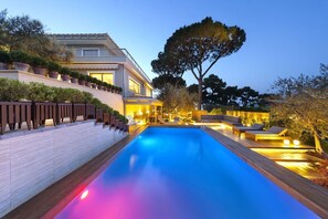 A modern poolside area with a wooden deck, lounge chairs, and a large tree.