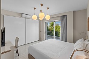 A modern hotel room with a large bed, a desk, and a balcony with a view of greenery.