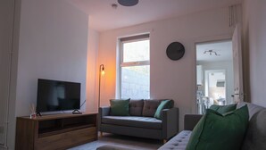 A living room with a TV on a wooden stand, a grey sofa with green cushions, and a window with a view of the outside. - 5BR House | Free Parking | Fast Wi-Fi | Sleeps 10 (Wolverhampton)