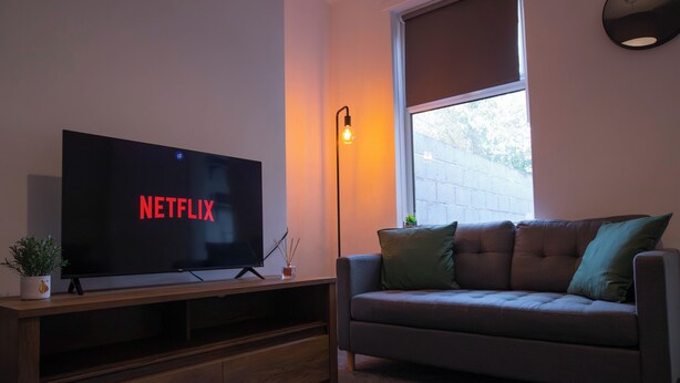 A living room with a flat-screen TV displaying "NETFLIX," a wooden entertainment unit, a sofa with cushions, a floor lamp, and a window with a view of a brick wall. - 5BR House | Free Parking | Fast Wi-Fi | Sleeps 10 (Wolverhampton)