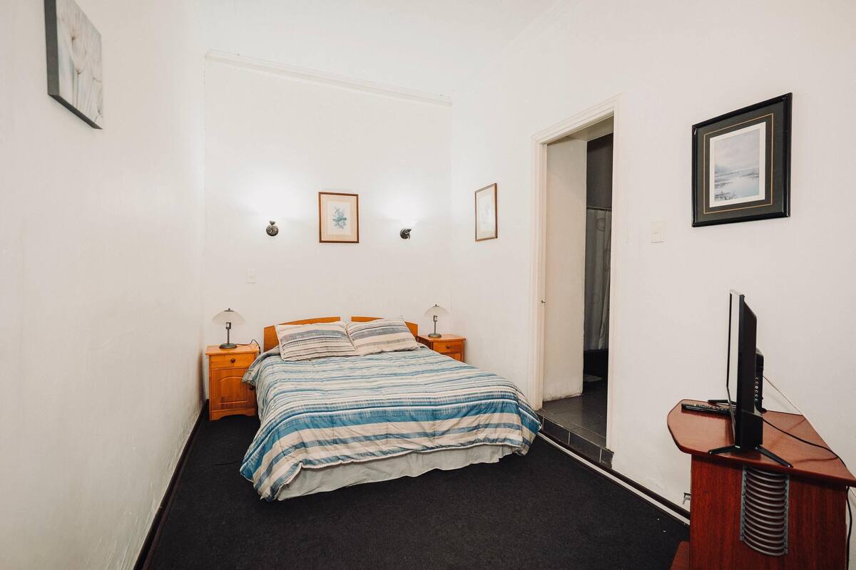 Un dormitorio con una cama, dos mesitas de noche con lámparas, un televisor sobre un mueble bajo y cuadros enmarcados en la pared.