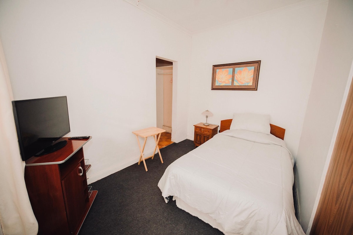 Habitación de hotel con una cama, un televisor sobre un mueble de madera, una mesita, una mesita de noche y un cuadro en la pared.