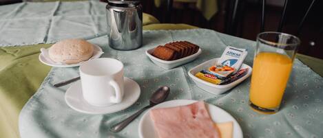 A breakfast setup with a coffee cup, muffin, ham slice, juice, and a packet of Azucar on a table with a green tablecloth.