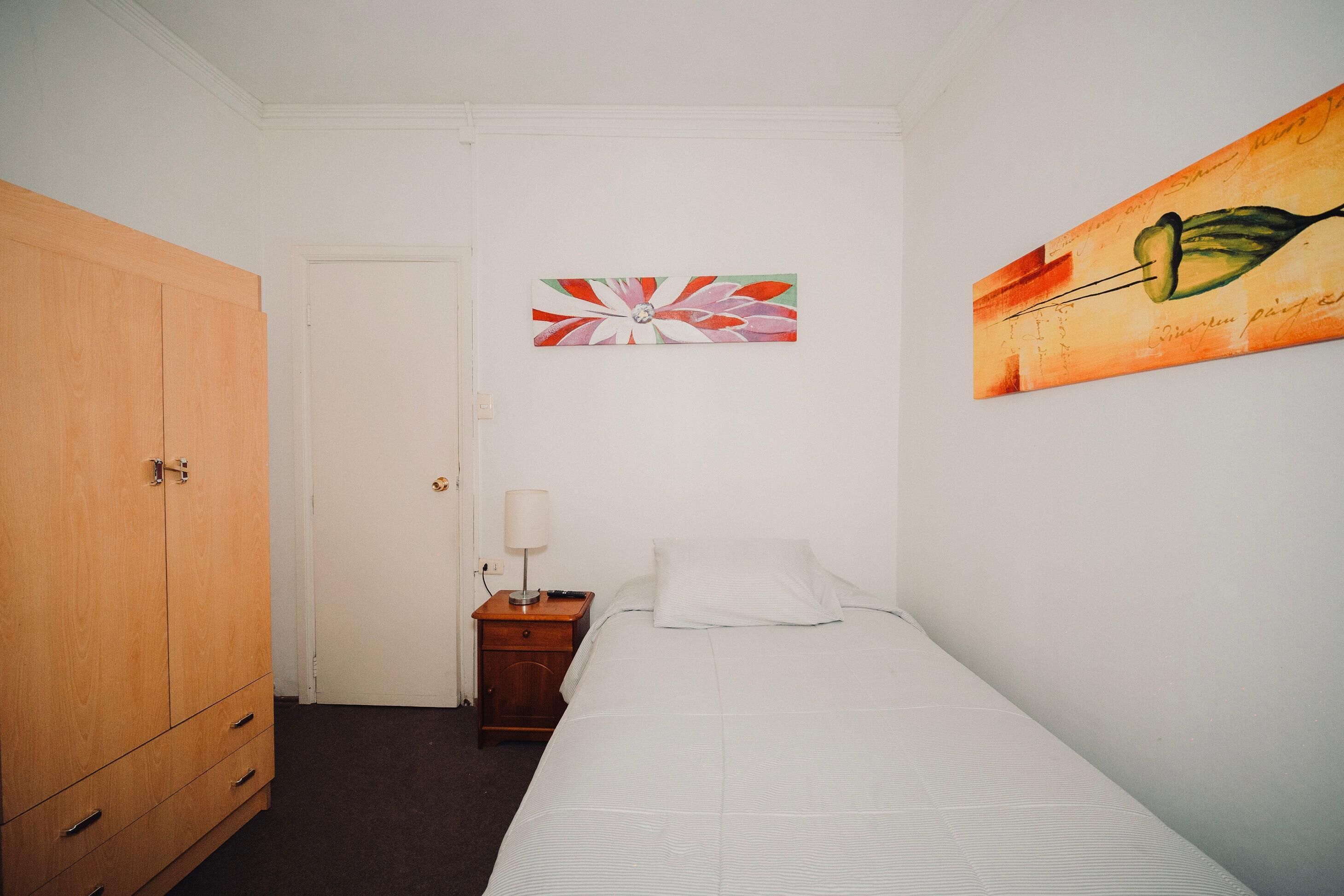 A bedroom with a single bed, a wooden wardrobe, a nightstand with a lamp, and two wall art pieces.