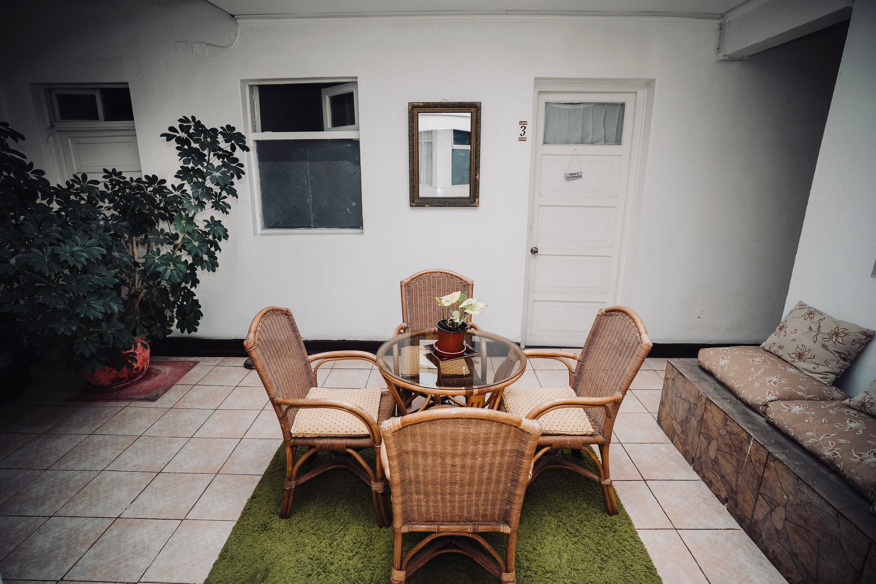 Un patio con una mesa redonda y cuatro sillas de mimbre, una planta en maceta y un sofá frente a una pared blanca.