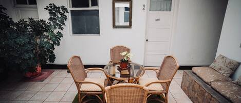 Un patio con una mesa redonda y cuatro sillas de mimbre, una planta en maceta y un sofá frente a una pared blanca.