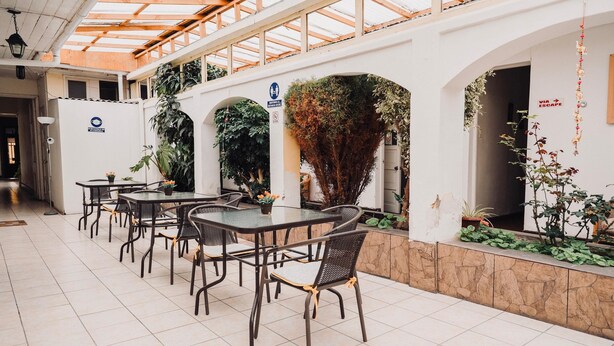 Un espacio de comedor al aire libre cubierto, con mesas y sillas, rodeado de plantas y un muro de piedra.