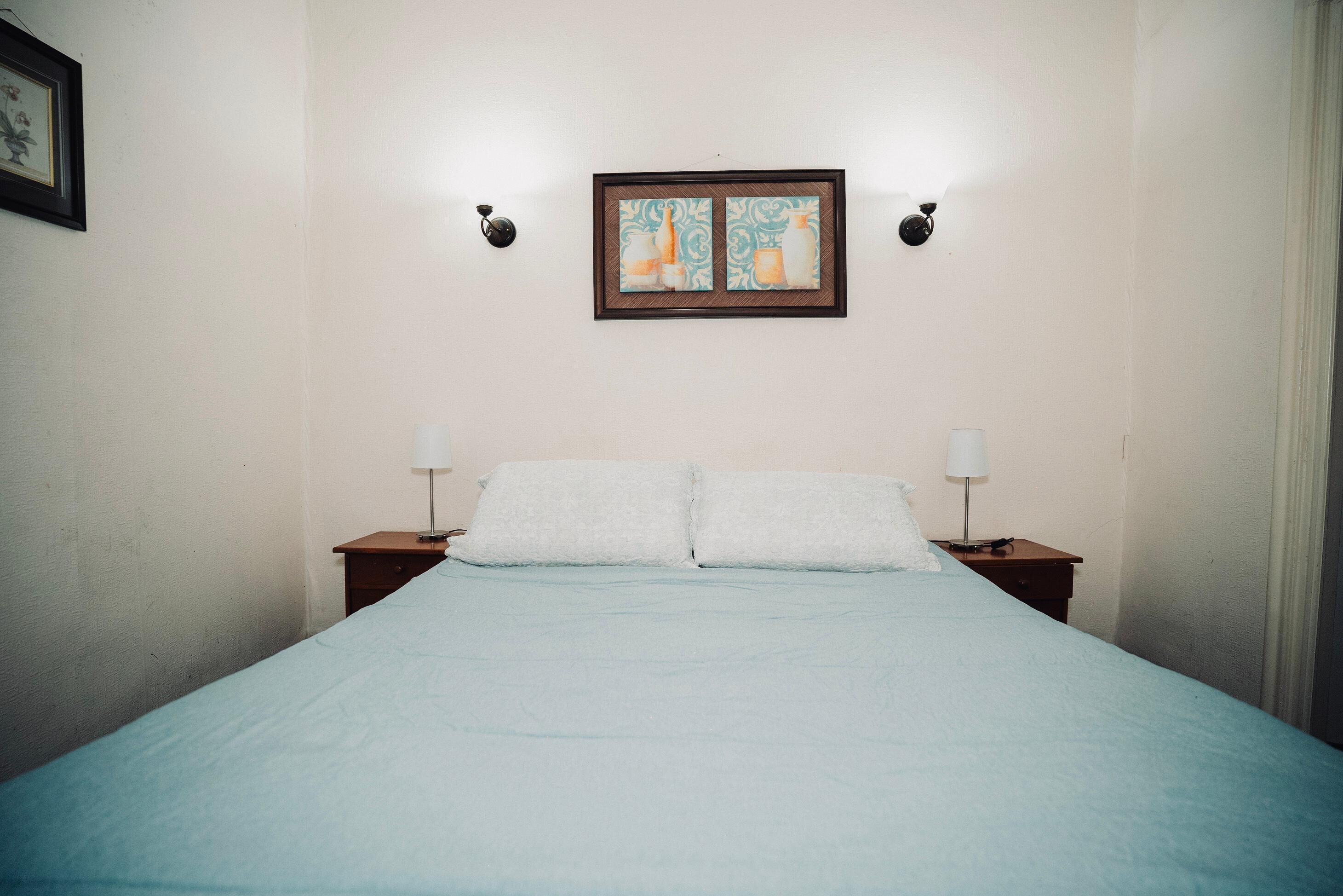 Una cama bien hecha con sábanas blancas, dos mesitas de noche con lámparas y una obra enmarcada sobre la cama.