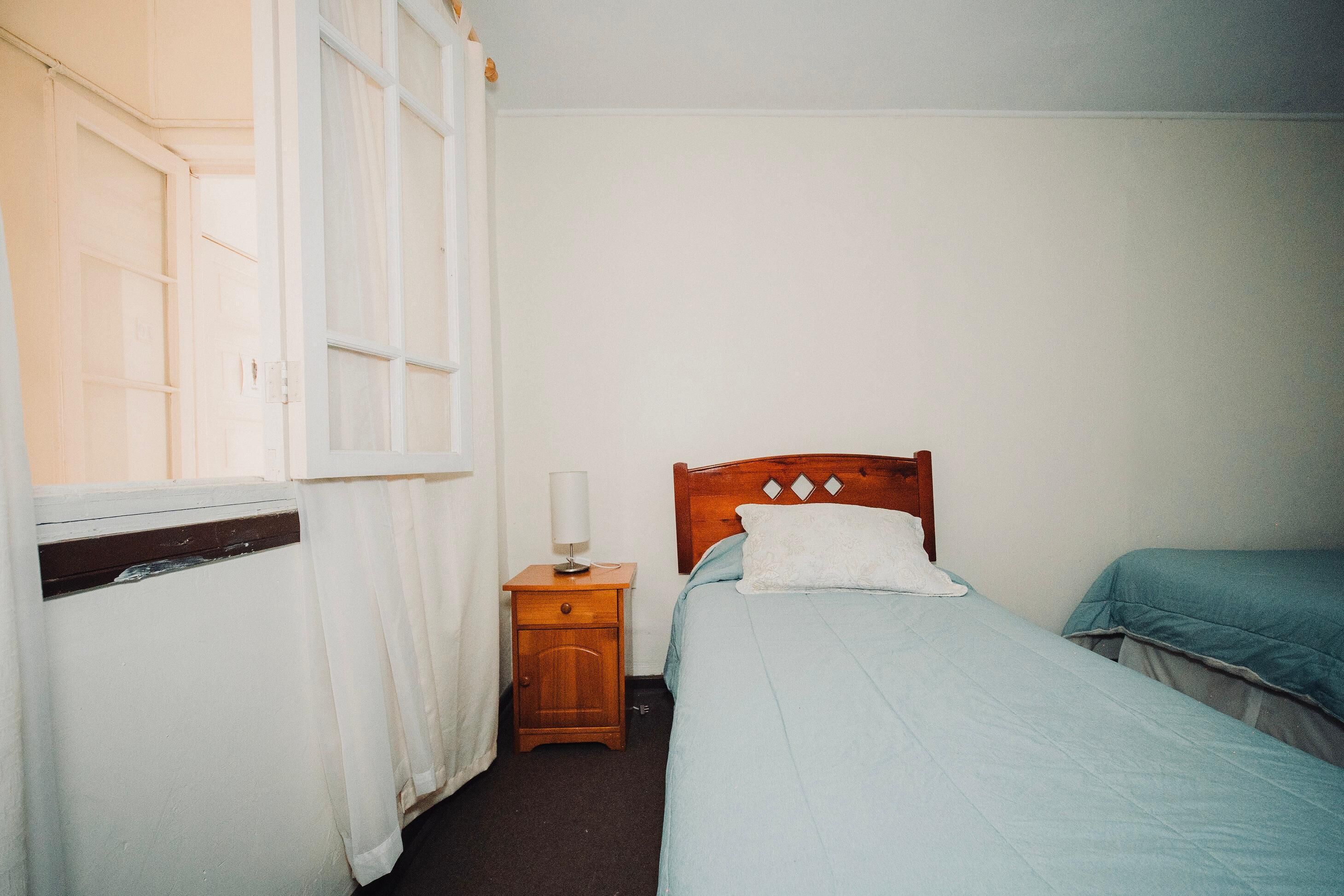 Quarto com uma cama de solteiro, mesa de cabeceira, janela com cortinas e radiador.
