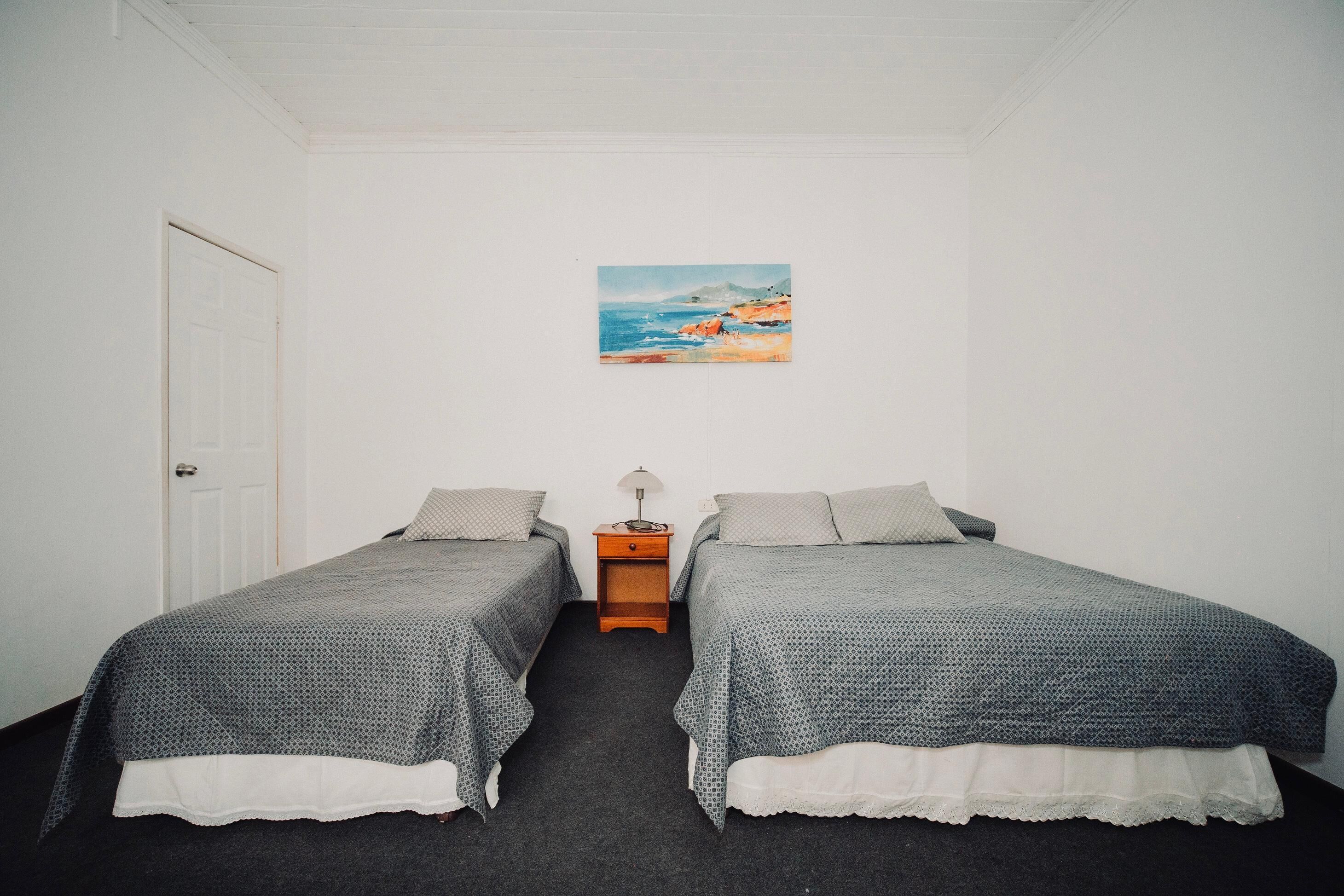 A room with two single beds, a small wooden nightstand, a white door, and a painting of a beach scene on the wall.