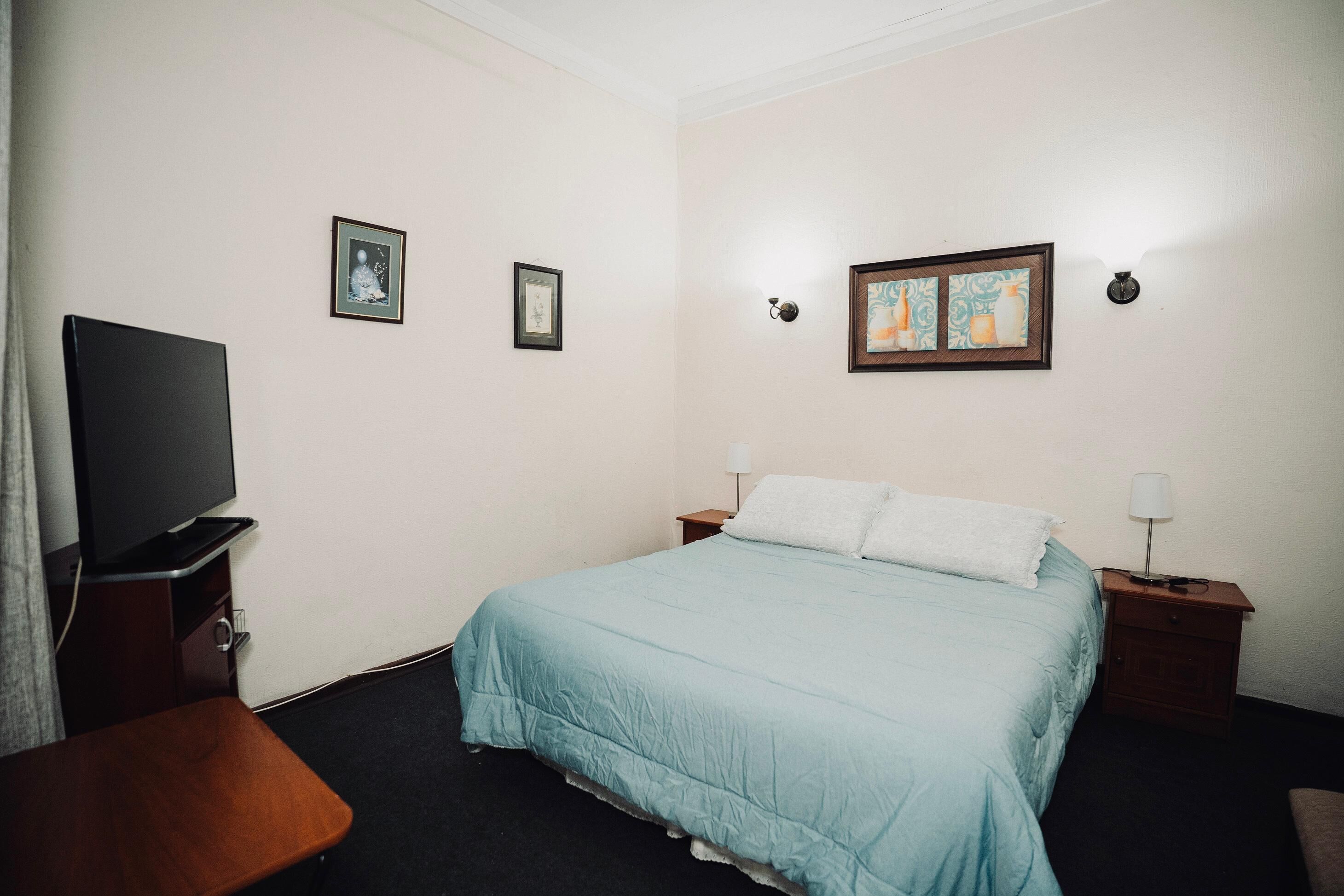A hotel room with a bed, a TV, a desk, and two framed pictures on the wall.