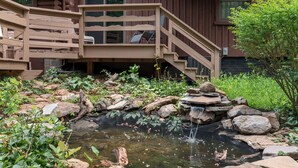 Une terrasse en bois avec des chaises, un petit étang avec des rochers et un escalier en bois menant à un bâtiment.