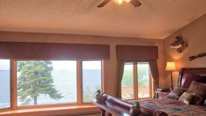 Quarto com uma grande janela com vista para um corpo de água, uma cama com colcha, uma mesa de cabeceira e um ventilador de teto.