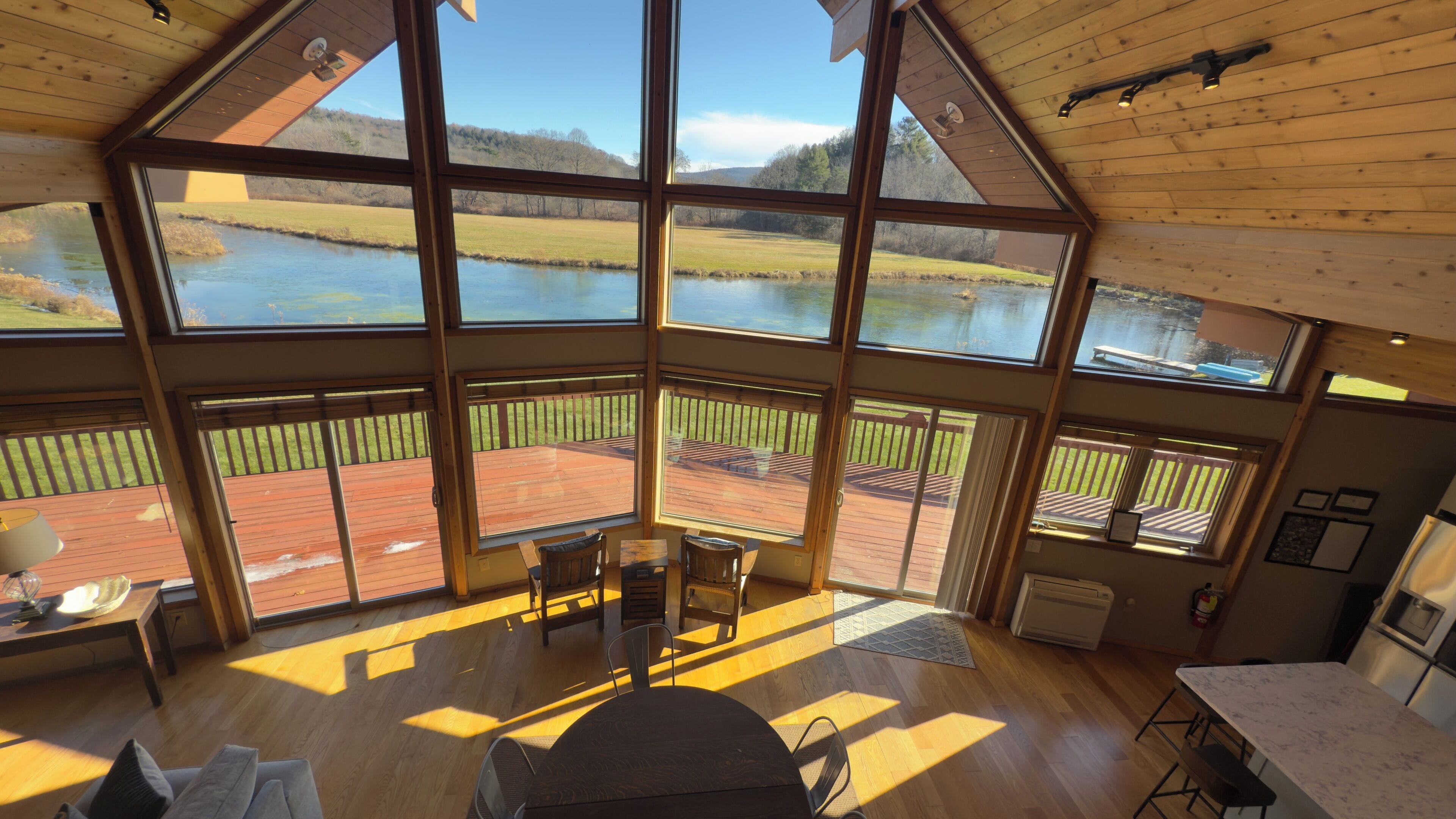 Une pièce spacieuse avec de grandes fenêtres donnant sur un lac, un plancher en bois et un coin repas comprenant une table et des chaises.