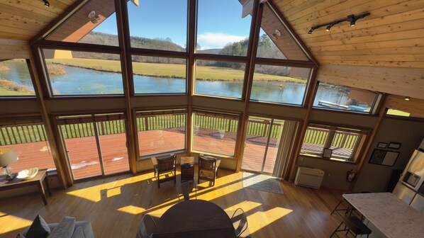 Quarto espaçoso com grandes janelas com vista para um lago, piso de madeira e área de jantar com mesa e cadeiras.