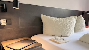 A neatly made bed with pillows, a laptop on a bedside table, and a book with glasses on it.