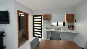 A compact kitchen with a gas stove, sink, and a small window. There is a dining table with chairs and a TV mounted on the wall. - Residencia Gran Coyula Orange 512 (Tonalá)