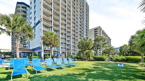 A high-rise hotel with multiple balconies, a well-maintained lawn with blue lounge chairs, and palm trees. - Our Newest Premium Ocean View Property - 2BR/2BA: Sleeps 8 - Top Amenities! (Myrtle Beach)