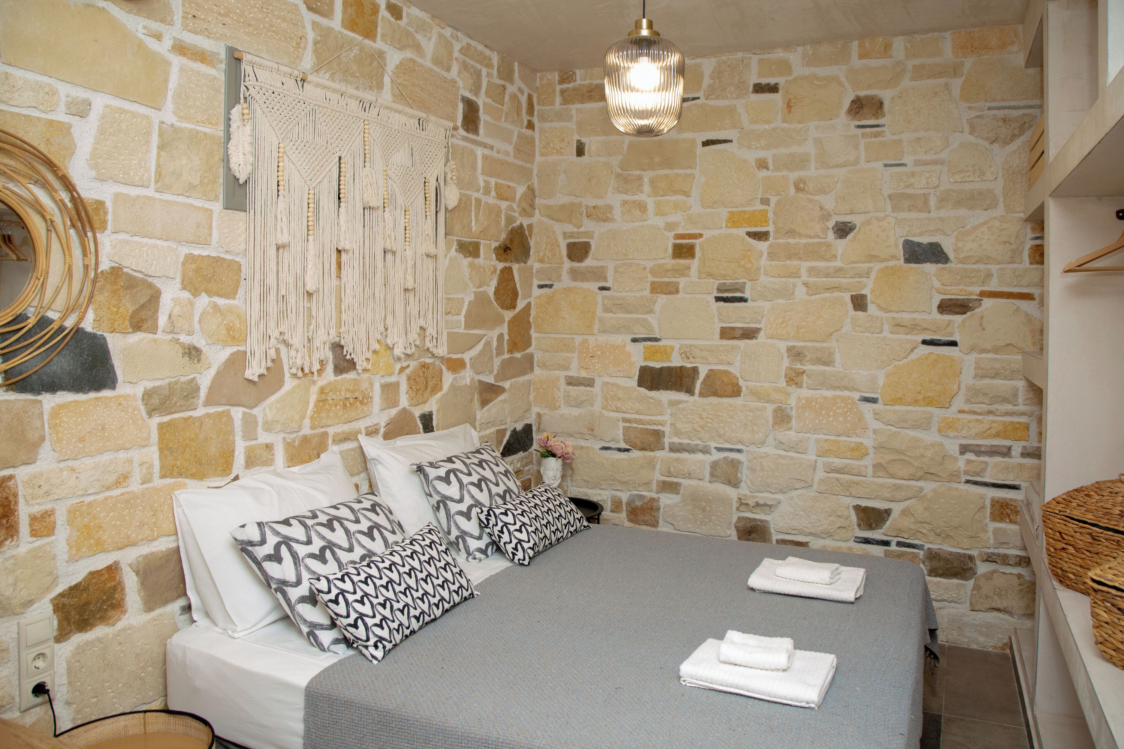 Quarto com paredes de pedra, uma cama, almofadas, um pendão de macramé e um lustre.