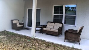 A covered patio area with a white exterior wall, two windows with blinds, and three wicker furniture pieces (two chairs and one sofa). - beautiful vacation home (Ocala)