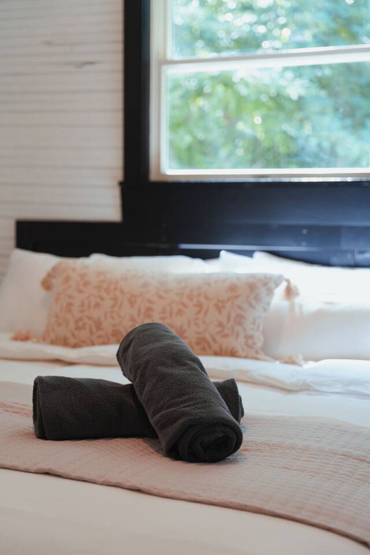 Una cama bien hecha con toallas enrolladas, una ventana con vistas a la vegetación y una almohada estampada.