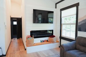 A living room with a fireplace, a television mounted above it, and a large window with a view of trees. - NEW! Historic Schoolhouse HotTub+FirePit+Wineries (Fauquier County)