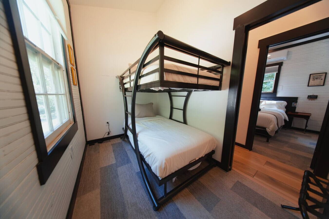 A bunk bed room with a window, carpeted floor, and a door leading to another room.