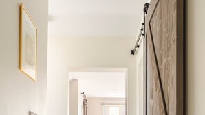 A hallway with a wooden door, a framed picture on the wall, and a view into another room.